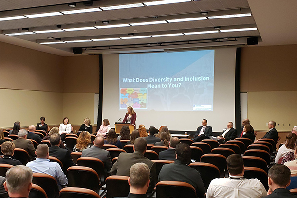 A conference room with audience and panelists members assembled in front of a screen that asks, "What does diversity and inclusion mean to you?"